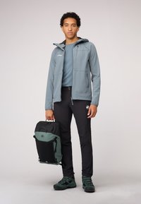 Lichte grijze jas, teal shirt, zwarte broek, groene accenten en een stevige zwart-groene rugzak, met robuuste zwarte wandelschoenen.