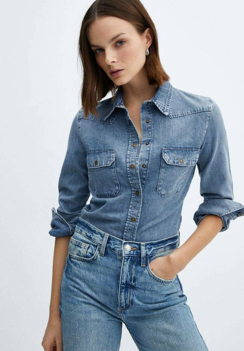 Hellblaue Denim-Bluse mit Knopfleiste und zwei Brusttaschen, kombiniert mit hoch taillierten Denim-Jeans. Der Stoff hat eine weiche, texturierte Oberfläche.