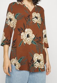 Top marrone con motivo floreale, scollatura a V e maniche a tre quarti. Presenta grandi fiori crema e foglie verdi. Tessuto morbido con una vestibilità morbida.