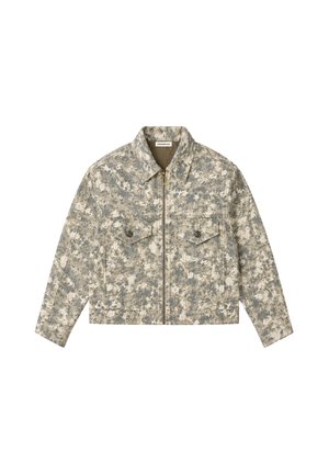 Ung mand iført camouflage lynlåstrøje over grå striktrøje med teksten "Berlin", står med hænderne i lommerne mod en ensfarvet baggrund.
