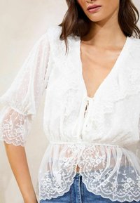 Blusa bianca con texture a pois, bordo in pizzo e scollatura arricciata. Il taglio morbido presenta una chiusura con nastro e maniche trasparenti.