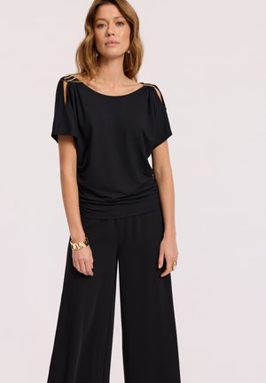 Czarny top bez rękawów z opadającymi rękawami i złotymi akcentami. W połączeniu z szerokimi czarnymi spodniami, prezentującymi gładką teksturę materiału.