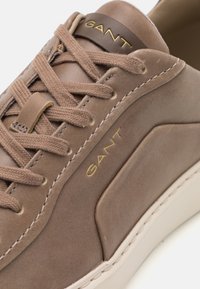 Braune Ledersneaker mit tonalen Schnürsenkeln, beigen Akzenten und einem goldenen Logo. Verfügt über ein schlankes Design und eine weiße Gummisohle.