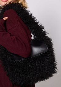 Kobieta w bordowym swetrze trzyma czarną, puszystą torebkę z ekologicznego futra z gładką skórzaną klapką na szarym tle.