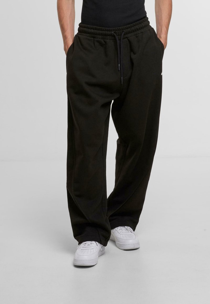 Black sweatpants with a relaxed fit, elastic waistband, and side pockets, paired with white sneakers. Smooth texture, no visible patterns.