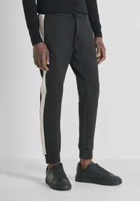 Zwarte joggers met een trekkoord in de taille, voorzien van crèmekleurige zijstrepen en enkelmanchetten. Gecombineerd met zwarte sneakers.