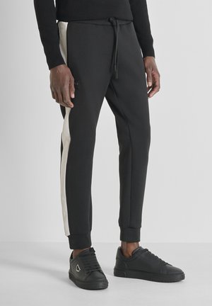 Black joggers with a drawstring waistband, featuring cream side stripes and ankle cuffs. Paired with black sneakers.