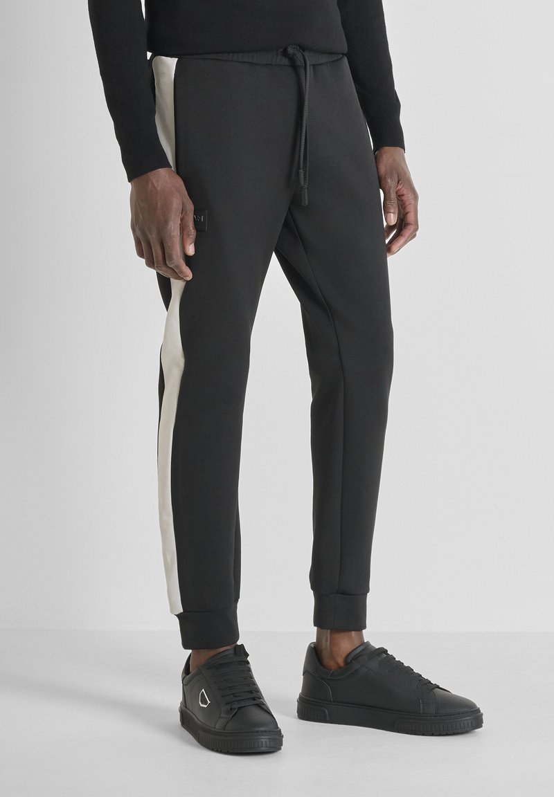 Zwarte joggers met een trekkoord in de taille, voorzien van crèmekleurige zijstrepen en enkelmanchetten. Gecombineerd met zwarte sneakers.