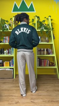 Πράσινο πανωφόρι varsity με λευκά γράμματα "BLKVIS" στην πλάτη, ριμπ μανσέτες και μέση, συνδυασμένο με μπεζ παντελόνια και λευκά παπούτσια.