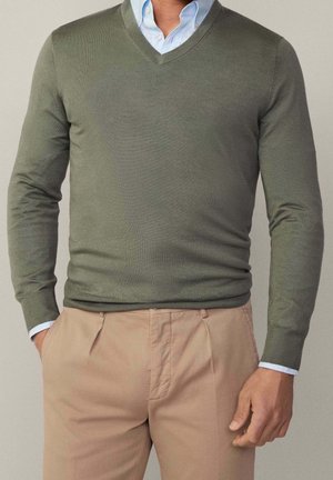 Mann trägt olivgrünen V-Ausschnitt-Pullover über hellblauem Hemd mit Kragen und beigefarbenen plissierten Hosen, steht mit einer Hand in der Tasche.