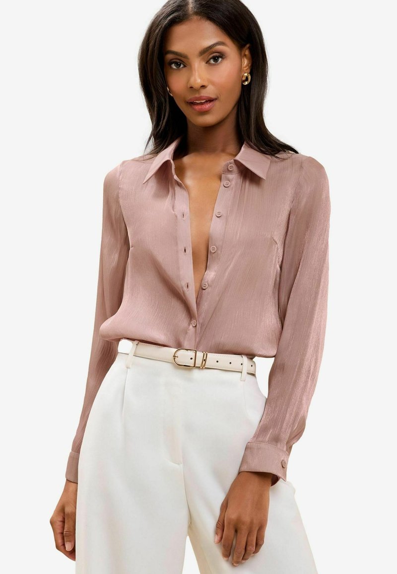 Helle pinke Bluse mit langen Ärmeln und Kragen, Knopfleiste und feiner Textur, kombiniert mit hoch geschnittenen weißen Hosen und einem Gürtel.