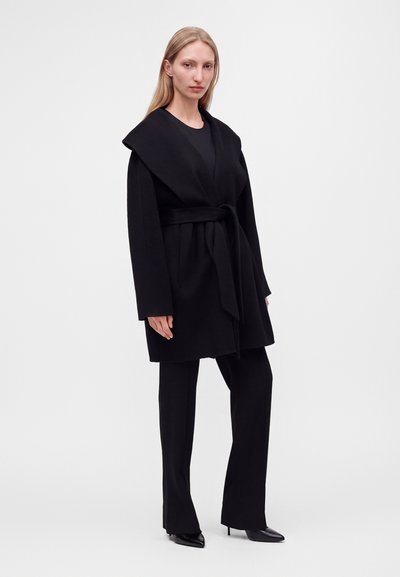 Femme debout portant un manteau noir ceinturé avec un large col, un pantalon noir et des chaussures à talons aiguilles noires sur un fond clair uni.