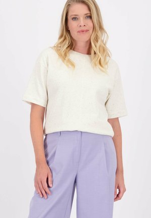 Blonde vrouw met losse golven, gekleed in een witte kortemouwen top en hoograndige lila wijde broek, staand voor een witte achtergrond.