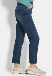 Jean en denim bleu foncé taille haute avec une coupe droite, cinq poches et des coutures visibles, associés à des baskets de sport de couleur claire.