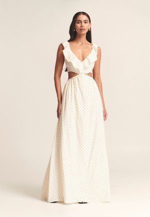 White maxi dress with black polka dots, ruffled neckline, open midsection, and flowing skirt. Made of lightweight fabric, designed for elegance.