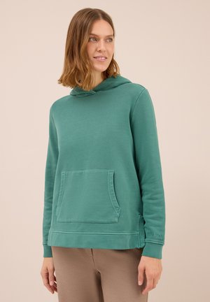 Frau in einem tealfarbenen Hoodie mit Fronttasche und beigen Hosen, die zur Seite schaut vor einem schlichten beigen Hintergrund.