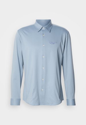 Ljusblå långärmad skjorta av mjukt tyg, med spetsig krage och knappar framtill, med en diskret broderad logga på bröstet.