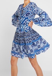 Modré vzorované šaty s kulatým výstřihem, dlouhými zvonovitými rukávy a nařaseným pasem, s různými odstíny modré a geometrickými vzory.