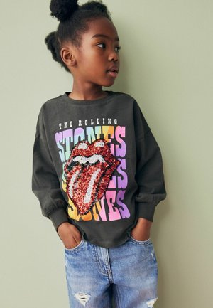 Sweatshirt grafic negru care prezintă un design mare cu buze roșii și albe, din paiete, pe un fundal de text colorat, asortat cu blugi albaștri și uzati.