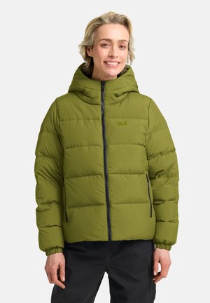 Lachend persoon met een olijfgroene pufferjas met capuchon en zwarte broek, staand tegen een effen lichte achtergrond.