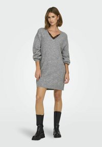 Robe pull grise avec un décolleté en V, dotée de manches surdimensionnées. Portée par-dessus un haut en dentelle noir, associée à des bottes noires à mi-mollet.