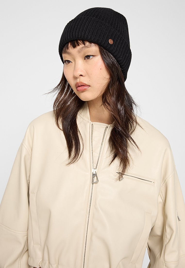 Czarna, prążkowana czapka beanie z małym, brązowym akcentem logotypu z metalu, noszona z kremową skórzaną kurtką bomberką z zamkiem błyskawicznym z przodu.