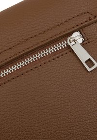 Pelle marrone con texture, dotata di una zip argento e un’etichetta per la tiratura. I punti di cucitura sono visibili lungo i bordi, evidenziando la resistenza del materiale.