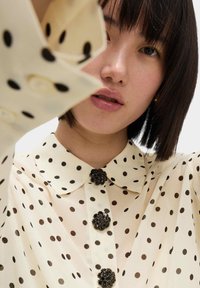Blusa di chiffon crema con pois neri, dotata di colletto e tre bottoni decorativi realizzati con ornamenti neri.