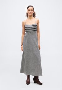 Γυναίκα που στέκεται φορώντας μαύρο και άσπρο καρό μίντι φόρεμα χωρίς μανίκια με λεπτές τιράντες και καφέ μπότες μέχρι τον αστράγαλο σε λευκό φόντο.