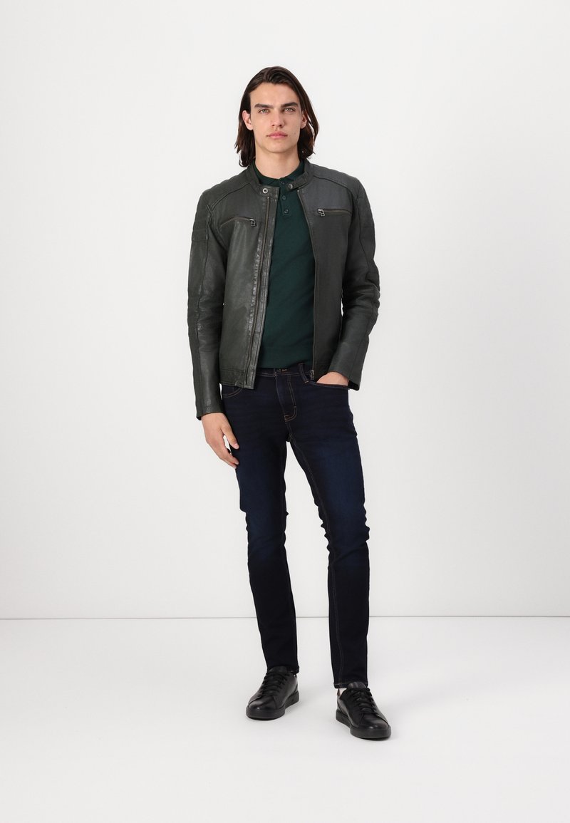 Veste en cuir vert foncé avec une coupe slim, dotée d'une fermeture éclair et de deux poches poitrine, associée à un polo vert foncé et à un jean bleu foncé.