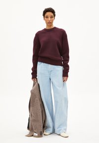 Bordeaux trui met lange mouwen en ribbelzoom, gecombineerd met lichtblauwe wijde jeans. Houdt een getextureerd bruin bovenstuk vast.