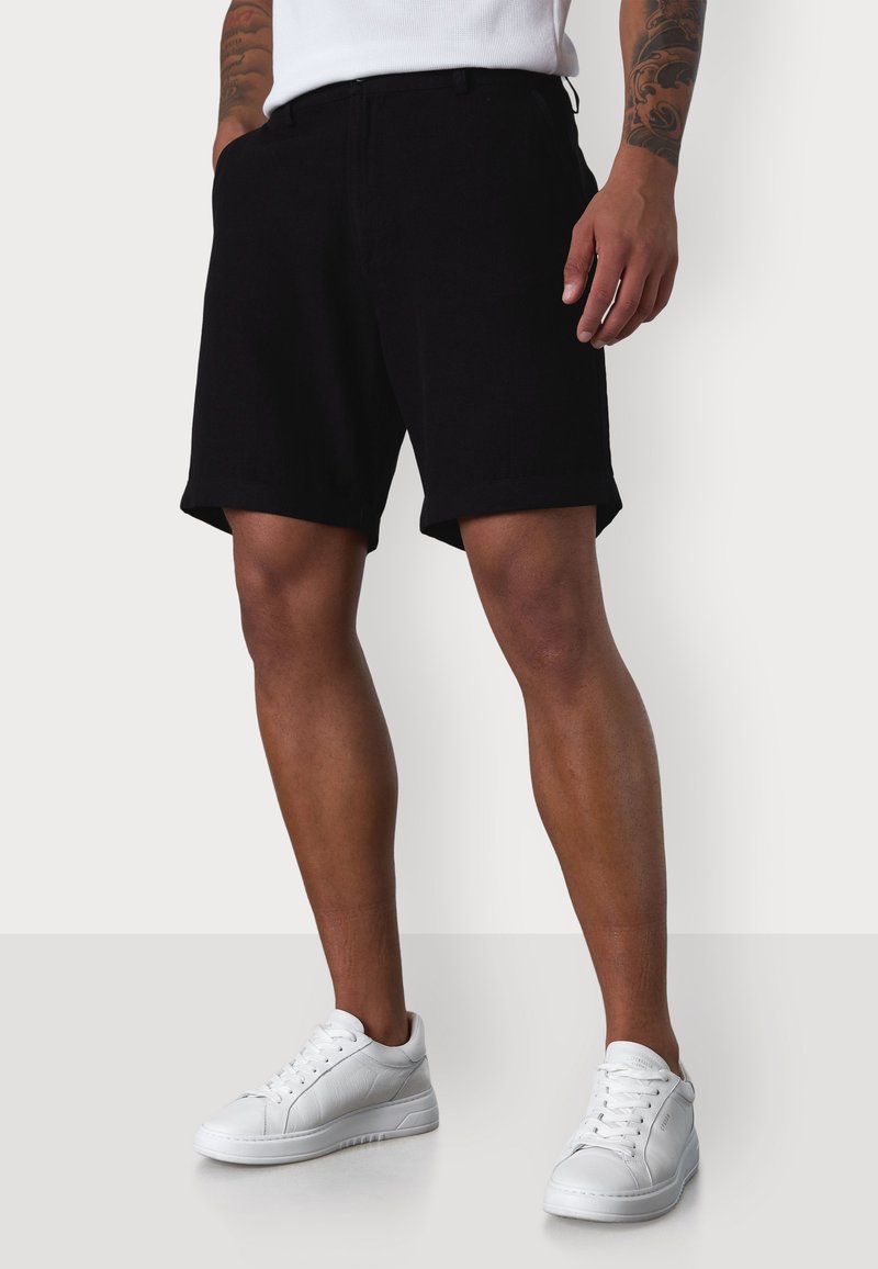 Schwarze Baumwollshorts mit flachem Vorderteil, umgeschlagenen Säumen und Seitentaschen, kombiniert mit weißen Sneakers. Schlichtes Design, keine sichtbaren Muster oder Akzente.