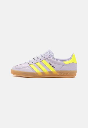 Sneaker Adidas Gazelle viola chiaro con strisce gialle e linguetta sul tallone, suola in gomma e lingua testurizzata su sfondo bianco.
