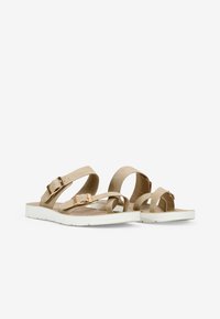 Sandales beiges avec une semelle blanche plate, dotées de brides croisées et d'une boucle en métal. Matériau lisse avec un design minimaliste.