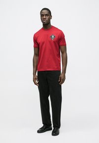 Rød bomulds t-shirt med rund hals og korte ærmer med et sort grafisk patch. Kombineret med sorte bukser med lige ben og sorte sneakers.