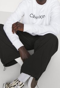 Witte sweatshirt met "Calvin Klein" logo, gecombineerd met losse zwarte broek. Schoeisel heeft een gestreept patroon. Eenvoudige, casual outfit.