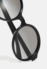 Óculos de sol redondos pretos com lentes escuras, armações grossas de plástico e hastes curvas. Detalhes na parte interna das hastes apresentam as especificações do modelo.