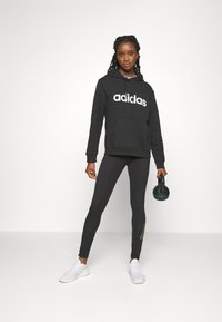 Črna kapuca s belim logotipom "adidas", črne legice in bele superge. Drži črn kettlebell in stoji na svetli podlagi.