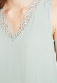 Haut sans manches vert clair avec un décolleté en V orné de délicates bordures en dentelle. Le tissu est doux avec une texture lisse, mettant en valeur les détails en dentelle.