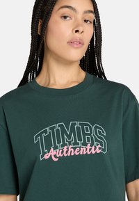 Dunkelgrünes Baumwoll-T-Shirt mit dem Schriftzug "TIMBS" in fettem weißen Text und "Authentic" in pinker Schreibschrift, kurzen Ärmeln und rundem Ausschnitt.