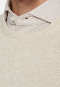 Nahaufnahme eines beigen Rundhals-Pullovers, der über einem hellbeigen Hemd mit Knöpfen getragen wird, wobei der oberste Knopf geschlossen ist.