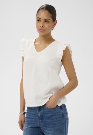 Femme aux cheveux foncés portant un chemisier blanc sans manches avec des épaules volantées et un jean bleu, main dans la poche, sur un fond uni.