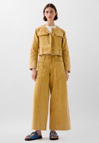 Giacca di jeans gialla con design accorciato e due tasche frontali, abbinata a pantaloni gialli a gamba larga. Il indossatore porta sandali blu.