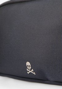 Bolso de tela negra texturizada con un pequeño emblema de calavera y huesos cruzados en la esquina inferior.