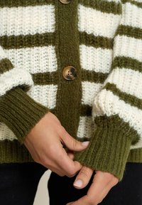 Gilet tricoté avec des rayures vert olive et blanches, de grands boutons et des poignets côtelés. Texture douce avec un design épais et chaud.