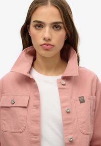 Giacca in cotone rosa con chiusura a bottoni, colletto ampio e taschino sul petto. Presenta accessori in argento e un'etichetta del marchio sulla tasca.