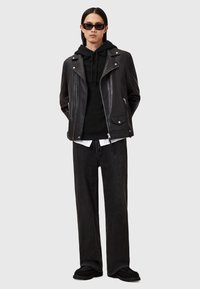 Veste de motard en cuir noir sur un hoodie noir, associée à un jean noir large et des chaussures noires. Des lunettes de soleil complètent le look.