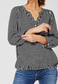 Gestreifte Bluse in Schwarz-Weiß mit Knopfleiste, langen Ärmeln, elastischem Saum und lockerer Passform. Getragen mit blauen Jeans.