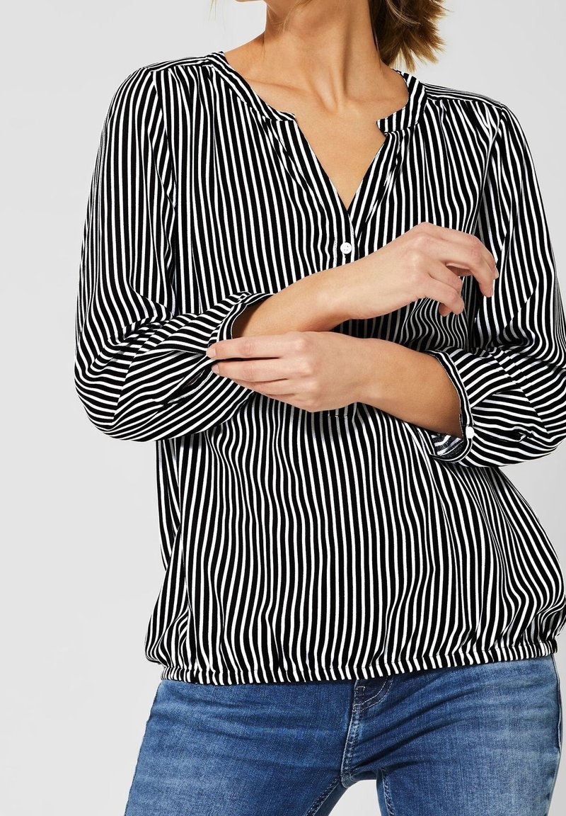 Gestreifte Bluse in Schwarz-Weiß mit Knopfleiste, langen Ärmeln, elastischem Saum und lockerer Passform. Getragen mit blauen Jeans.