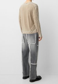 Béžová košile s dlouhým rukávem a šedé džíny s náplastovým vzorem a třepenými okraji; texturovaná látka, volný střih a černé boty.